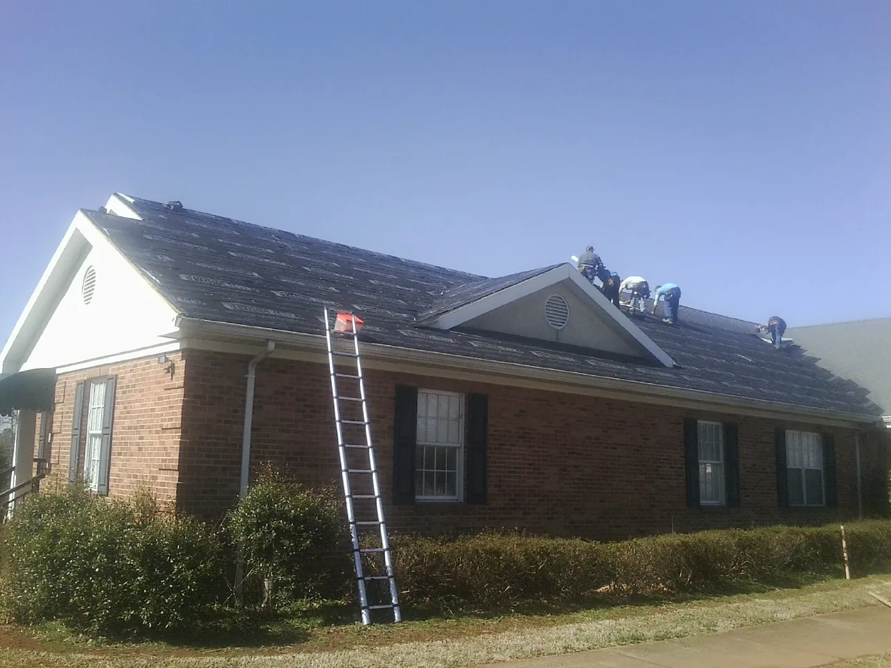 Crew tearing shingles off a brick ranch roof with a ladder set — Emergency Roof Repair work by our crew in Picture Rocks