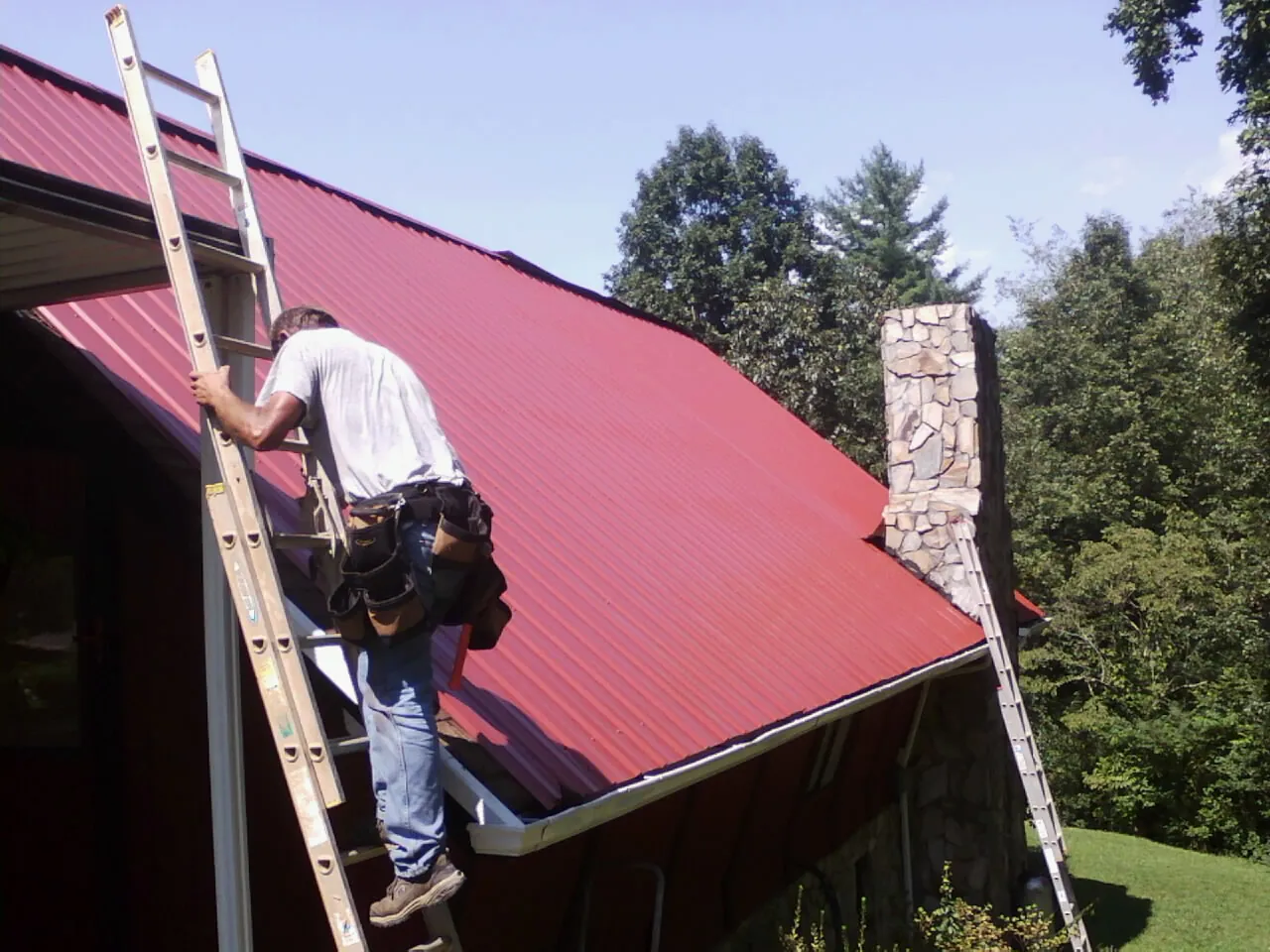Roofer on a ladder beside a red metal roof and stone chimney — Soffit & Fascia Repair by Roofing Brothers in Picture Rocks